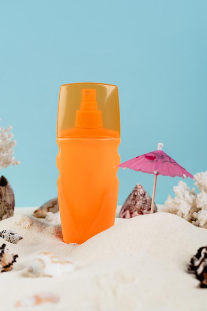 oranžová sprej láhev opalovacího krému v blízkosti mušlí na písku izolované na modré - Fotografie, Obrázek