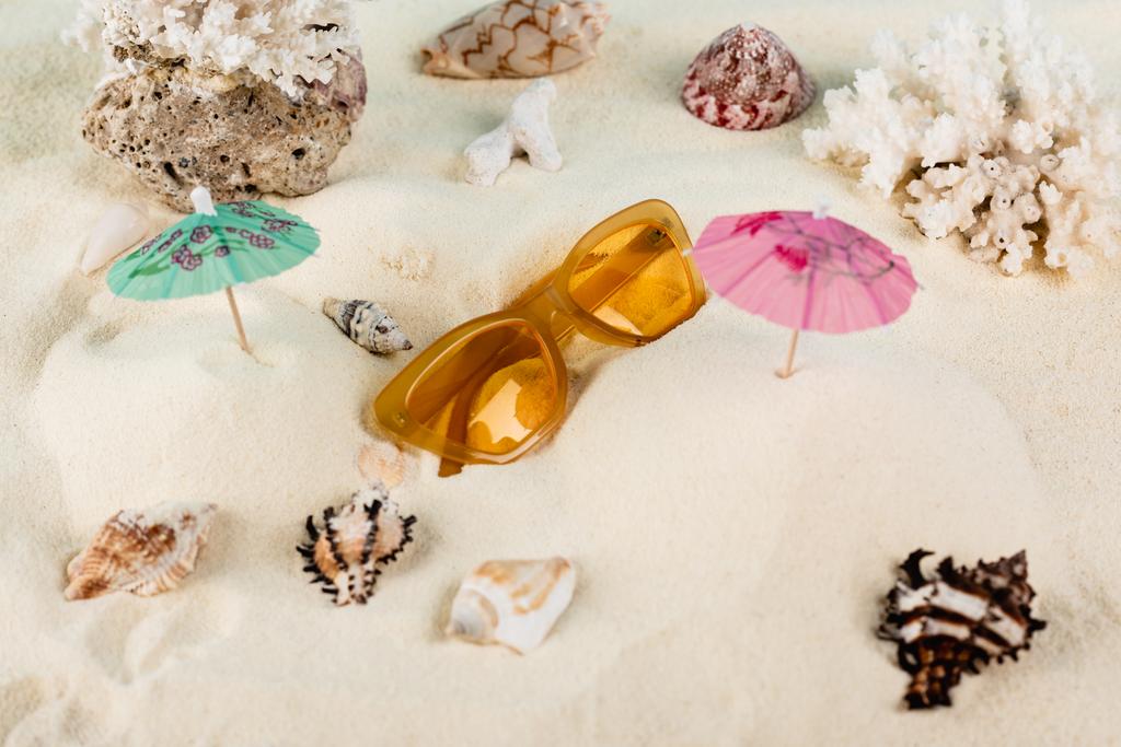 top view of orange sunglasses near seashells and cocktail umbrellas on sand - Φωτογραφία, εικόνα