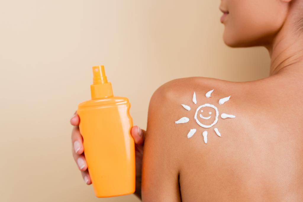 cropped view of woman with image of smiling sun on shoulder holding sunscreen isolated on beige - Foto, Imagem