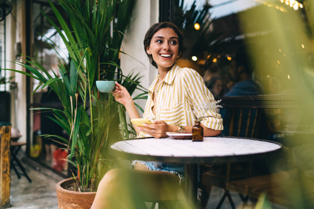 Усміхнена молода жінка в повсякденному одязі п'є чашку кави і використовує мобільний телефон, сидячи за невеликим круглим столом і насолоджуючись часом у вуличному кафе
 - Фото, зображення