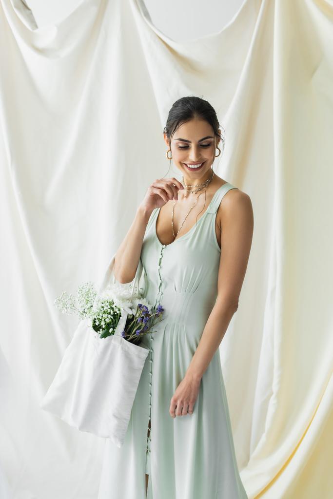 mujer alegre sosteniendo bolso de comprador de tela con flores en blanco - Foto, imagen