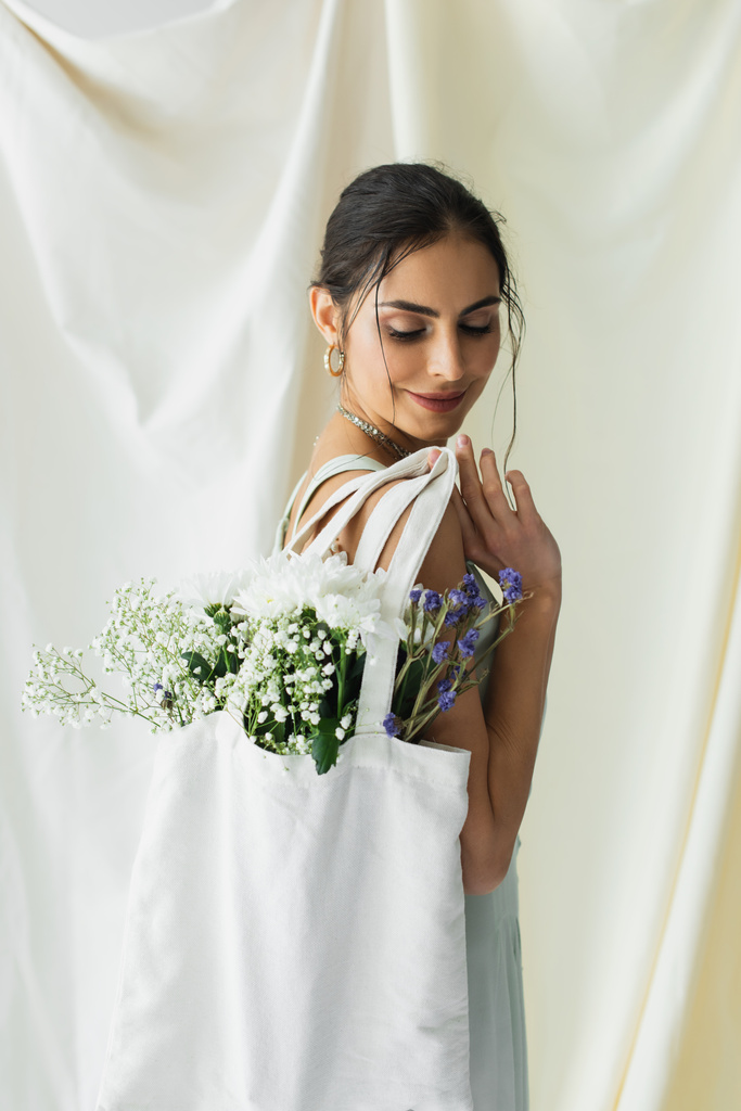 vrolijke vrouw houden stof shopper tas met bloemen op wit - Foto, afbeelding