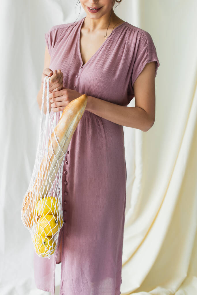 bijgesneden uitzicht van gelukkige vrouw met herbruikbare string zak met citroenen en stokbrood op wit  - Foto, afbeelding