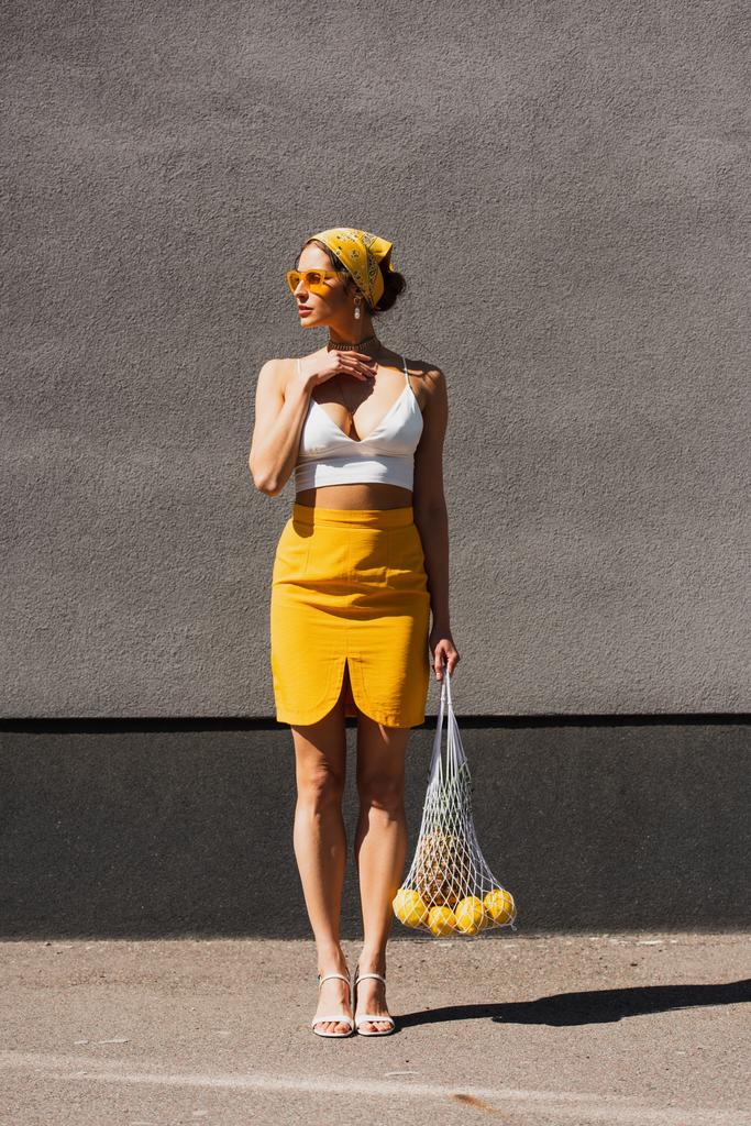 pleine longueur de femme élégante dans des lunettes de soleil et foulard jaune tenant sac à ficelle avec des fruits près du mur en béton  - Photo, image