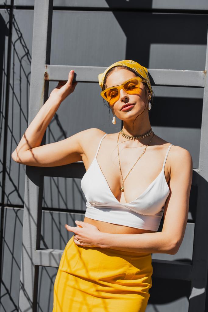 μοντέρνα γυναίκα με γυαλιά ηλίου και κίτρινη μαντίλα που ποζάρει κοντά σε τσιμεντένιο τοίχο  - Φωτογραφία, εικόνα