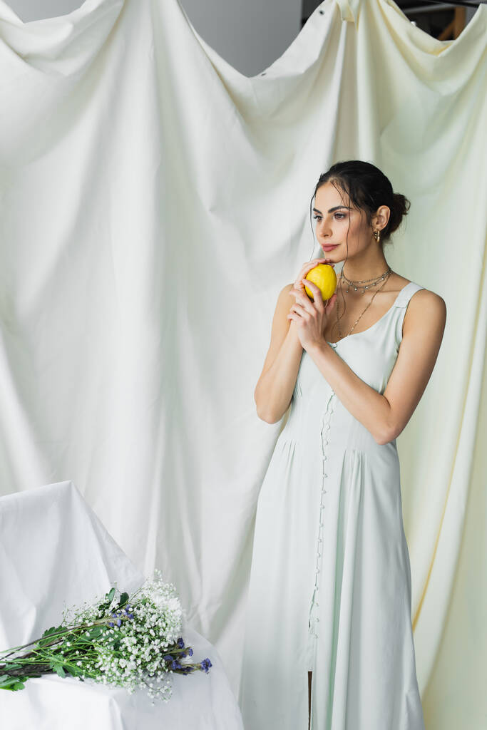 мечтательная женщина в платье с лимоном возле букета цветов на белом - Фото, изображение