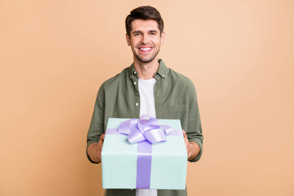 Photo de charmant jeune homme heureux donner boîte cadeau vous sourire célébrer vacances isolé sur fond de couleur beige - Photo, image