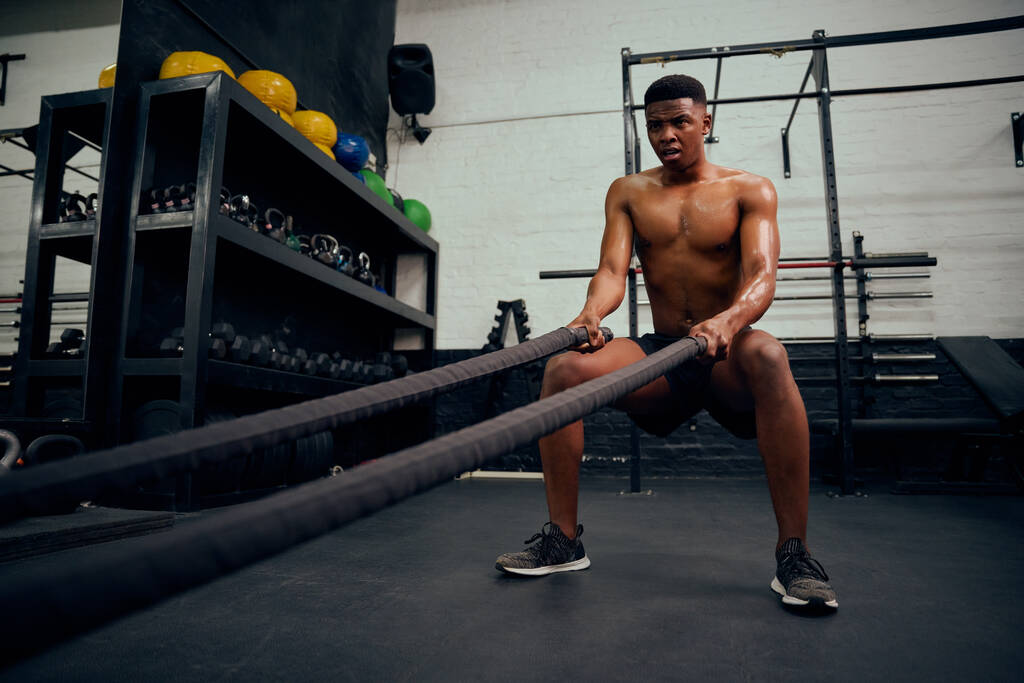 Giovane maschio afroamericano che si allena dentro una palestra. Topless personal trainer maschile utilizzando corda per rafforzare le braccia. Foto di alta qualità - Foto, immagini