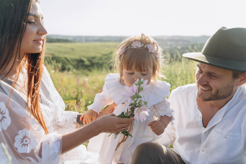 Feliz familia joven pasar tiempo juntos en la naturaleza, hermosa mujer en vestido de guipur blanco con el marido y su hija sentada en el campo - Foto, Imagen