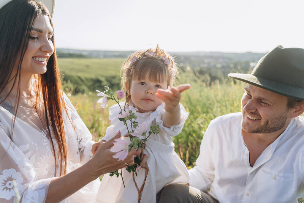 Onnellinen nuori perhe viettää aikaa yhdessä luonnossa, kaunis nainen valkoisessa guipure-mekossa miehensä ja tyttärensä kanssa, jotka istuvat kentällä. - Valokuva, kuva