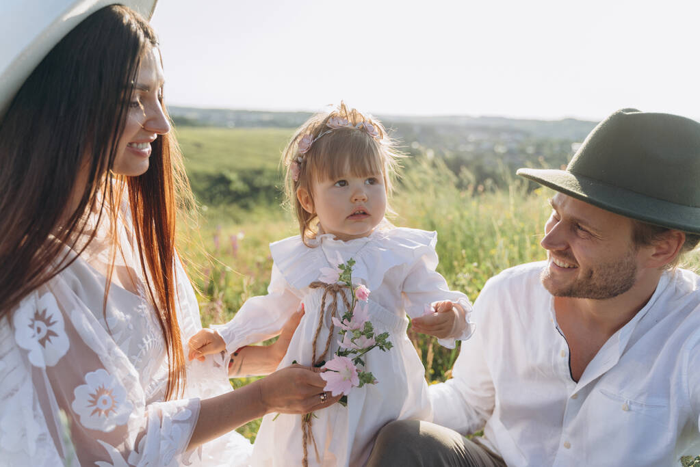 Onnellinen nuori perhe viettää aikaa yhdessä luonnossa, kaunis nainen valkoisessa guipure-mekossa miehensä ja tyttärensä kanssa, jotka istuvat kentällä. - Valokuva, kuva