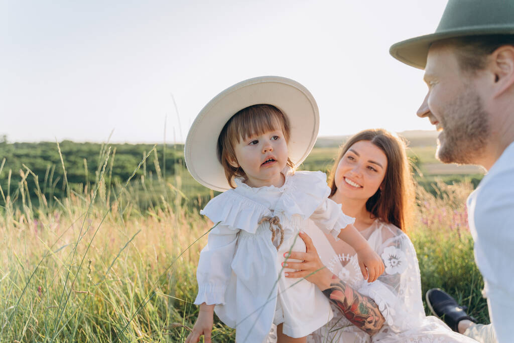 Šťastná mladá rodina tráví čas spolu v přírodě, krásná žena v bílých guipure šaty s manželem a jejich dcerou sedí v terénu - Fotografie, Obrázek