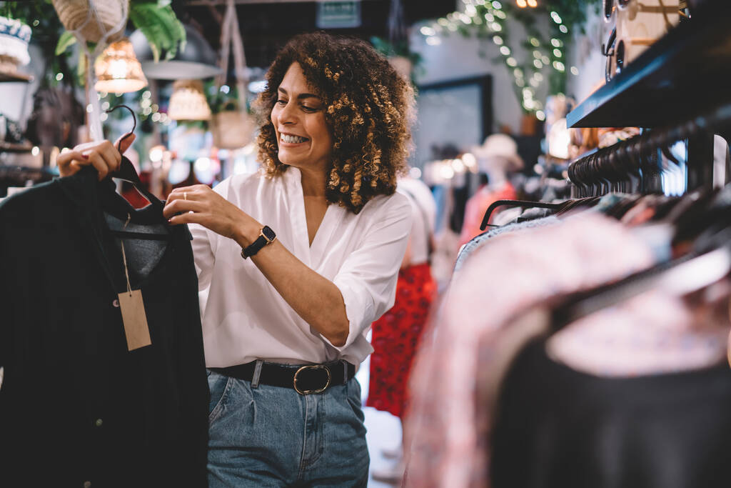 Cheerful female standing near hangers with brand clothes and laughing during Black Friday shopping with discounts, happy hipster girl enjoying pastime for update her wardrobe spending day in boutique - 写真・画像