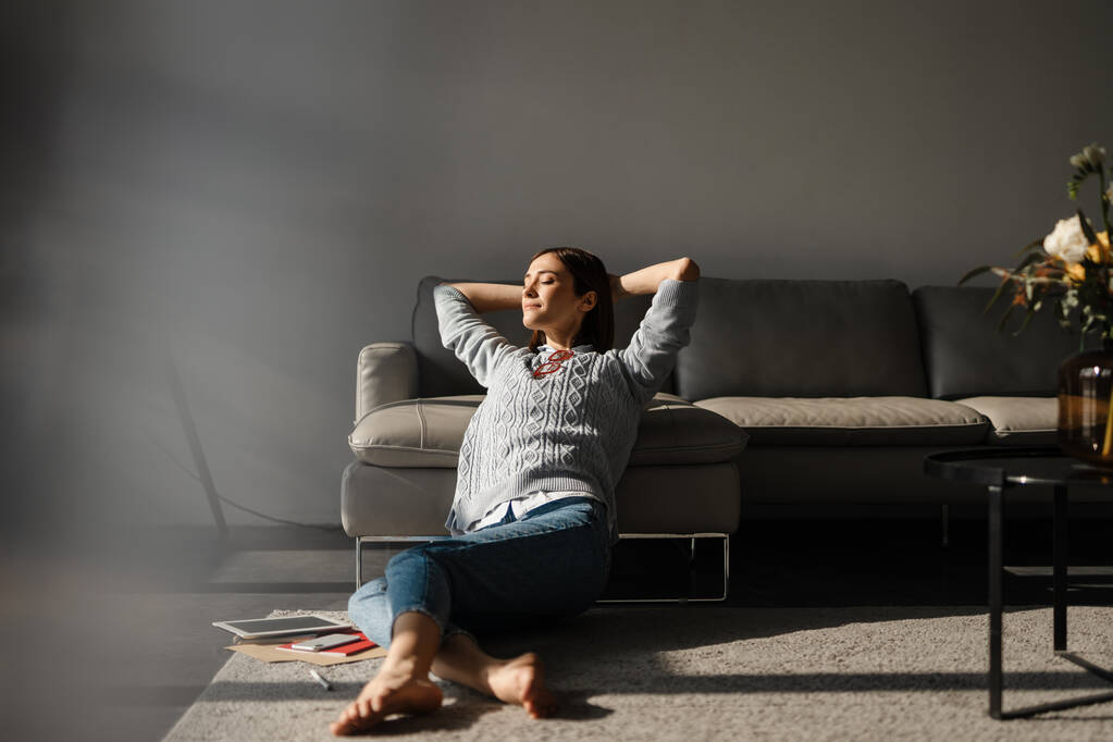 Weiße junge Frau sitzt zu Hause auf dem Boden, die Hände hinter seinem Hals verschränkt - Foto, Bild