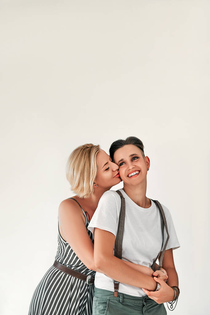 Portrait of a smiling two lesbian women, one kisses another on the cheek on white background. Vertical photo. - Foto, Imagem