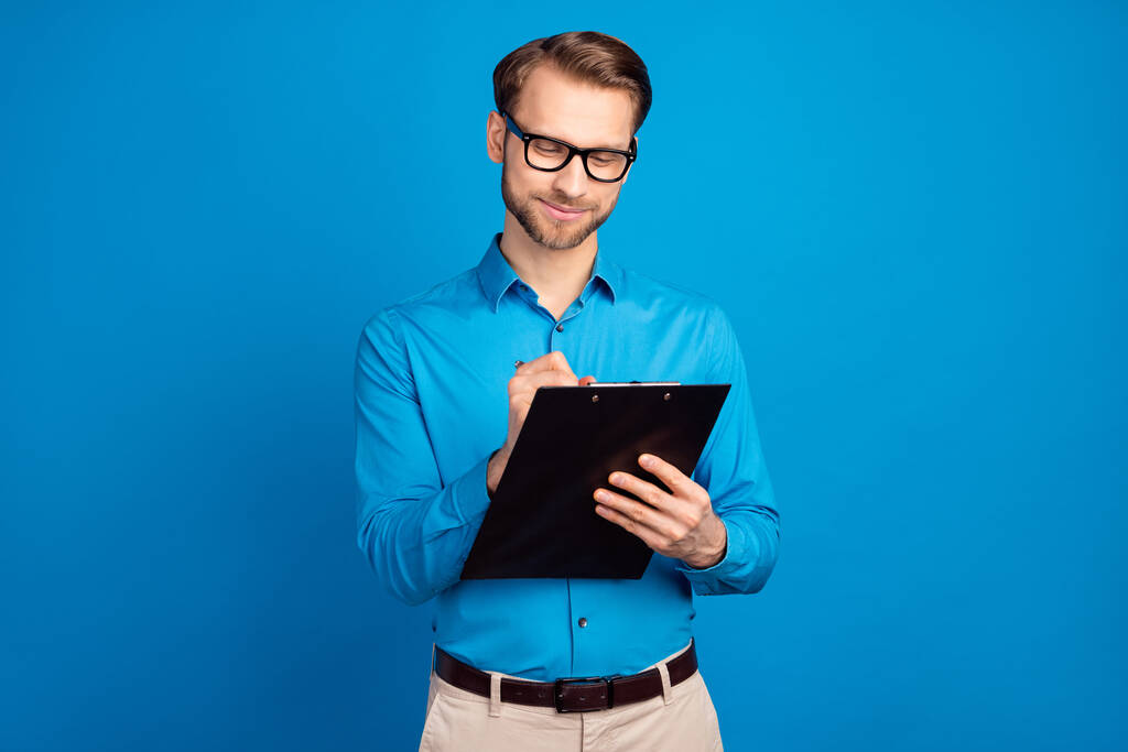 Фото молодого бизнесмена счастливой позитивной улыбкой написать заметки план буфера обмена изолированы на синем фоне цвета - Фото, изображение