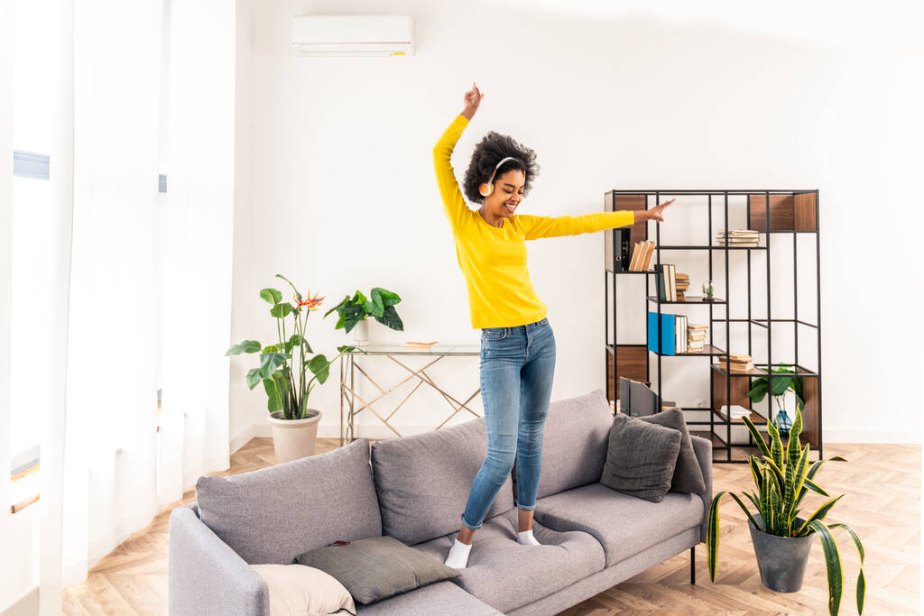 Ευτυχισμένη γυναίκα που ακούει μουσική, πηδάει και χορεύει στον καναπέ στο σπίτι - Φωτογραφία, εικόνα
