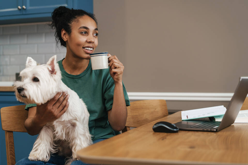 家のキッチンで犬と座っている間にコーヒーを飲む黒人女性の笑顔 - 写真・画像