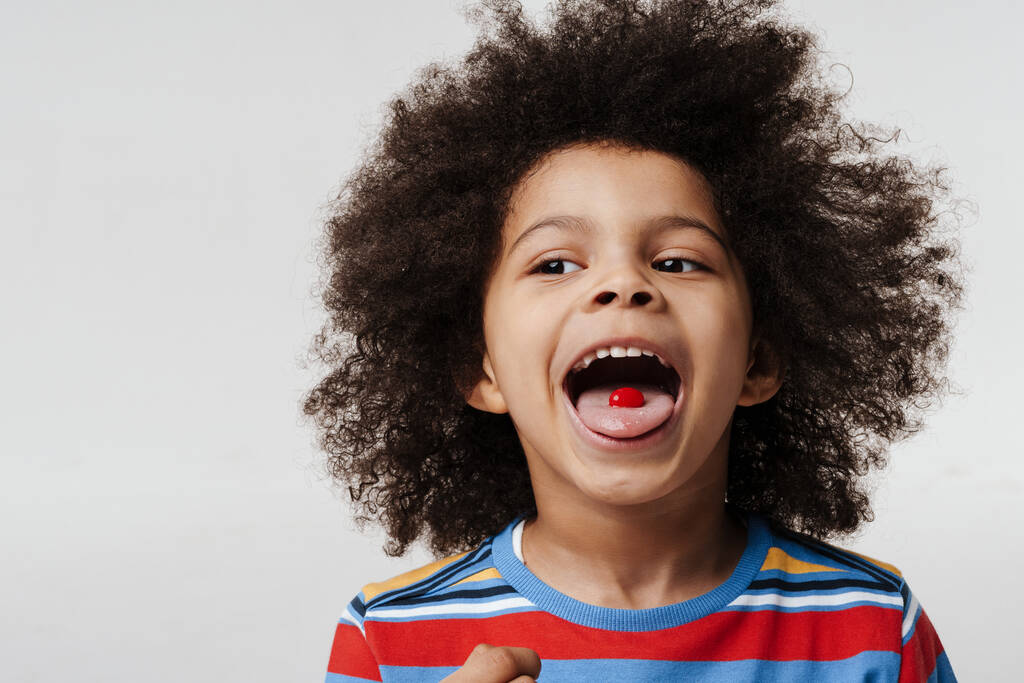 Großaufnahme eines glücklichen afrikanischen Vorschulkindes, der ein T-Shirt über der weißen Wand trägt und seine Zunge mit einem Bonbon herausstreckt - Foto, Bild