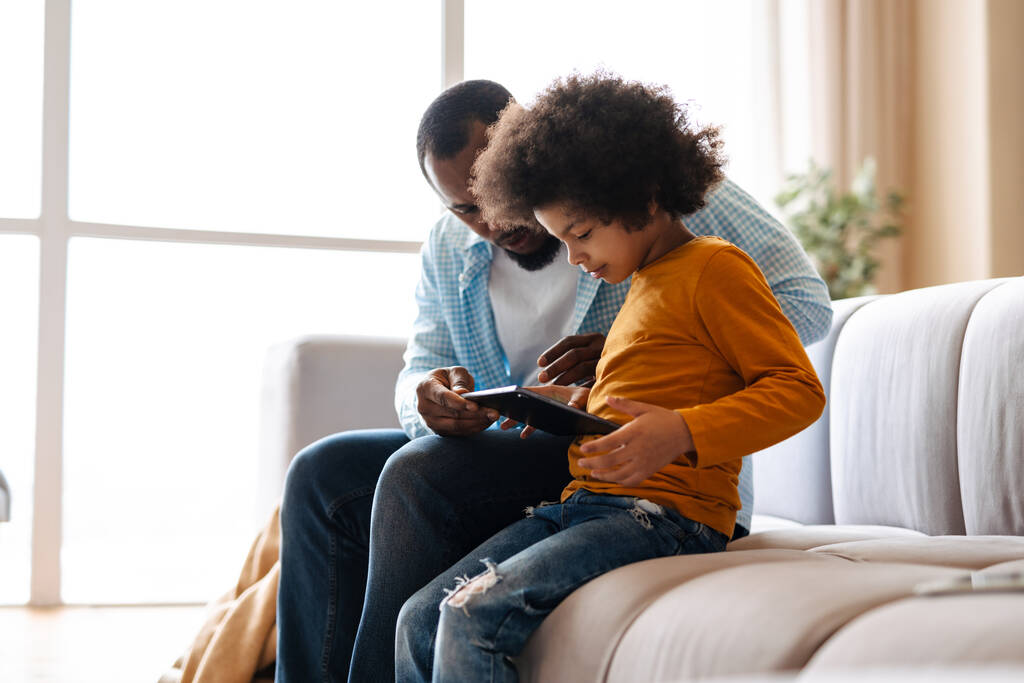 Μαύρος πατέρας και γιος χρησιμοποιώντας το tablet υπολογιστή, ενώ κάθεται στον καναπέ στο σπίτι - Φωτογραφία, εικόνα