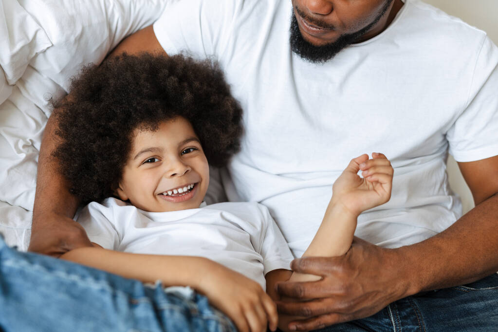 Μαύρος πατέρας και γιος γελούν ενώ κάνουν πλάκα στο κρεβάτι στο σπίτι - Φωτογραφία, εικόνα