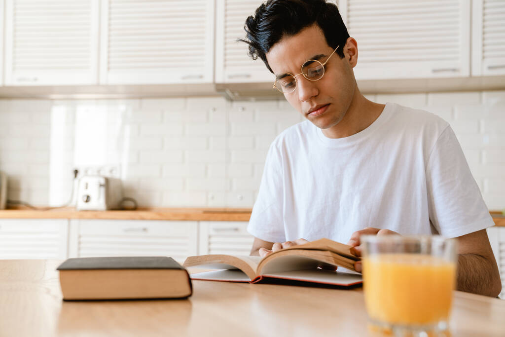 A fiatal spanyol férfi könyvet olvas, miközben otthon a konyhában reggelizik, az asztalnál ülve. - Fotó, kép