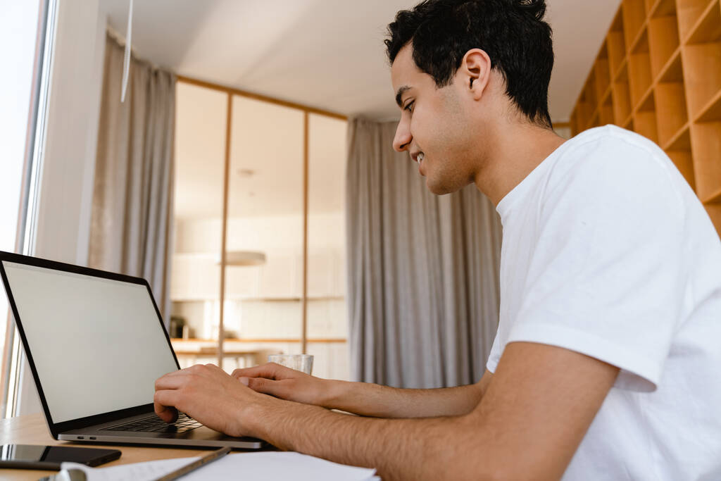 Hombre hispano joven pensativo que estudia en la computadora portátil mientras está sentado en el escritorio en casa - Foto, Imagen