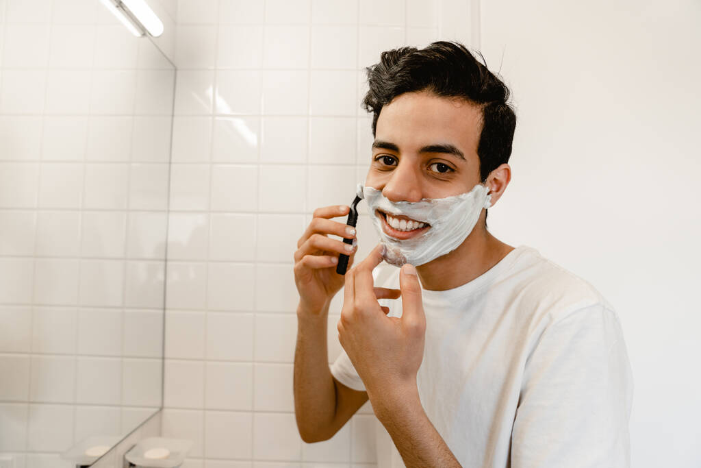 glimlachende jonge latino man scheren zijn gezicht staan voor de spiegel binnen - Foto, afbeelding