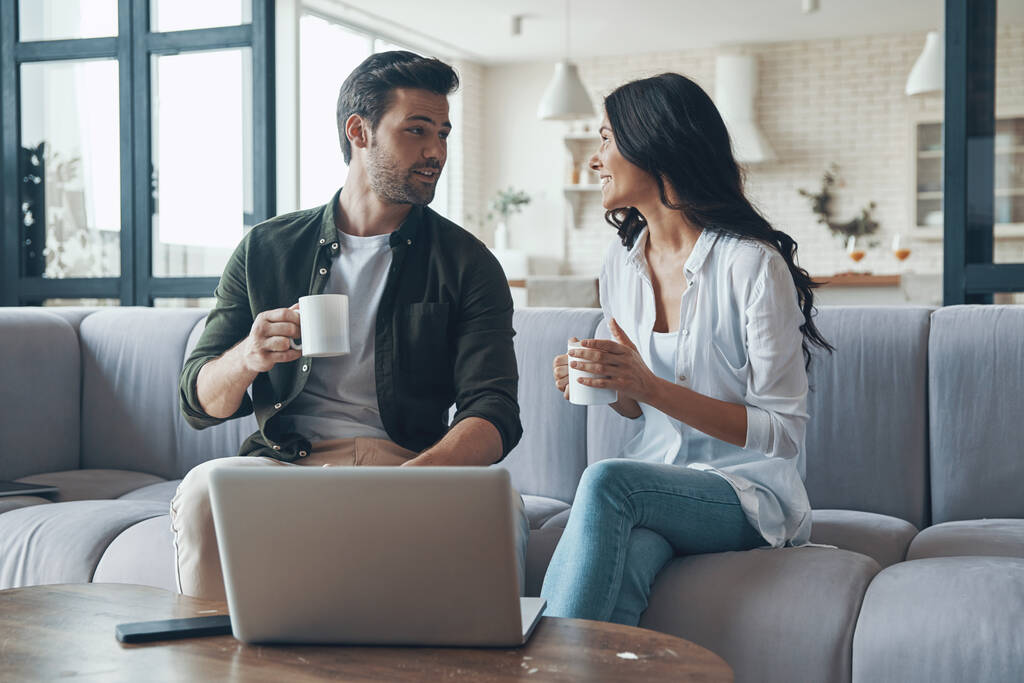 Piękna młoda para pijąca kawę i patrząca na laptopa siedząc na kanapie w domu - Zdjęcie, obraz