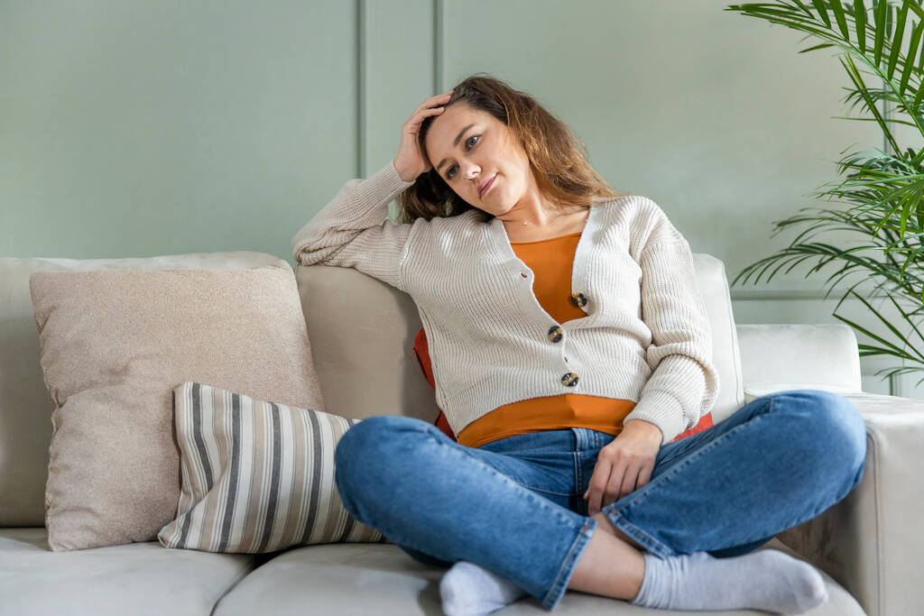 Νεαρή γυναίκα ξαπλωμένη στο σπίτι, στο σαλόνι, καθισμένη στον καναπέ. Νιώθει λυπημένη και ανήσυχη που υποφέρει από κατάθλιψη στην ψυχική υγεία. Προβλήματα και ραγισμένη καρδιά έννοια. - Φωτογραφία, εικόνα
