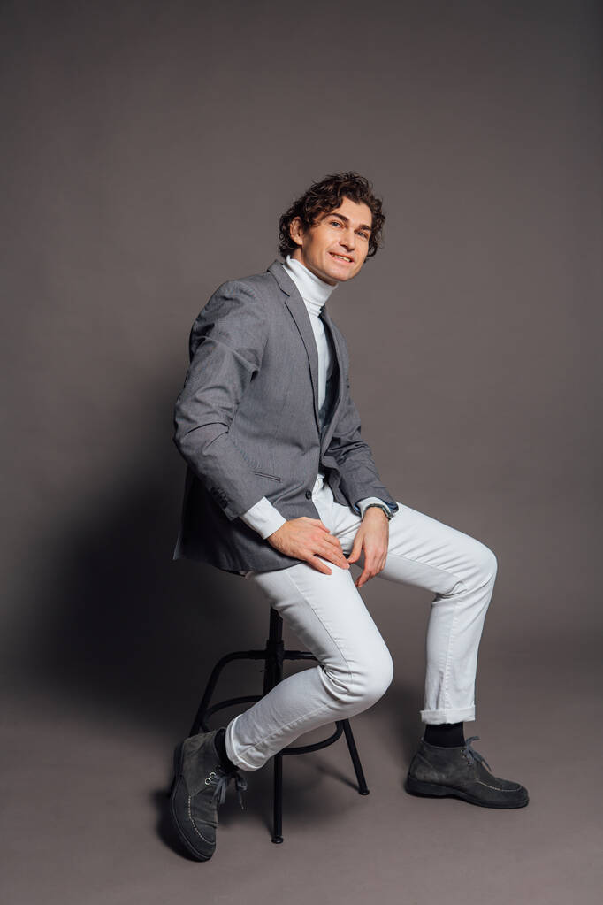 Retrato de moda de um homem bonito alto vestido com gola alta branca, jeans brancos e casaco cinza sentado na cadeira e posando no fundo cinza - Foto, Imagem
