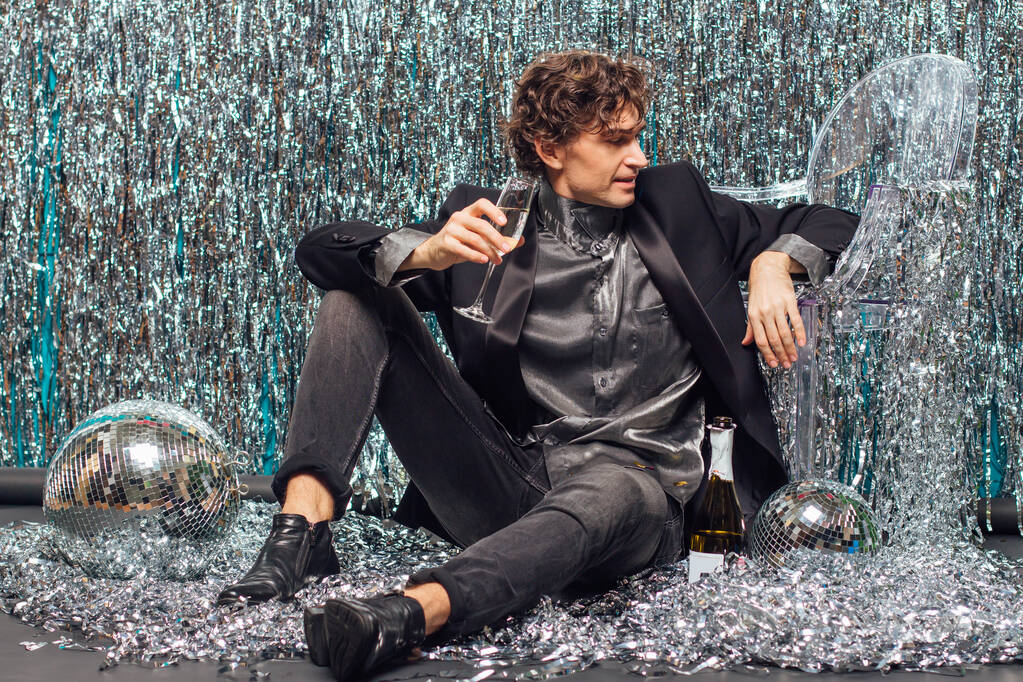 Un hombre alto y atractivo sentado en el suelo sobre el fondo brillante y la bola disco espejo sosteniendo una copa de champán. Concepto del partido. - Foto, Imagen