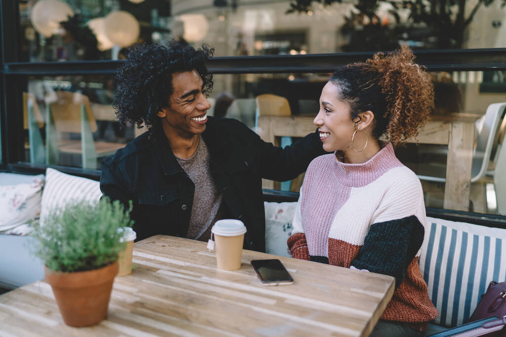 Joyeux petit ami multiethnique et petite amie souriant franchement les uns aux autres tout en étant assis à petite table avec café pour aller dans un café moderne - Photo, image