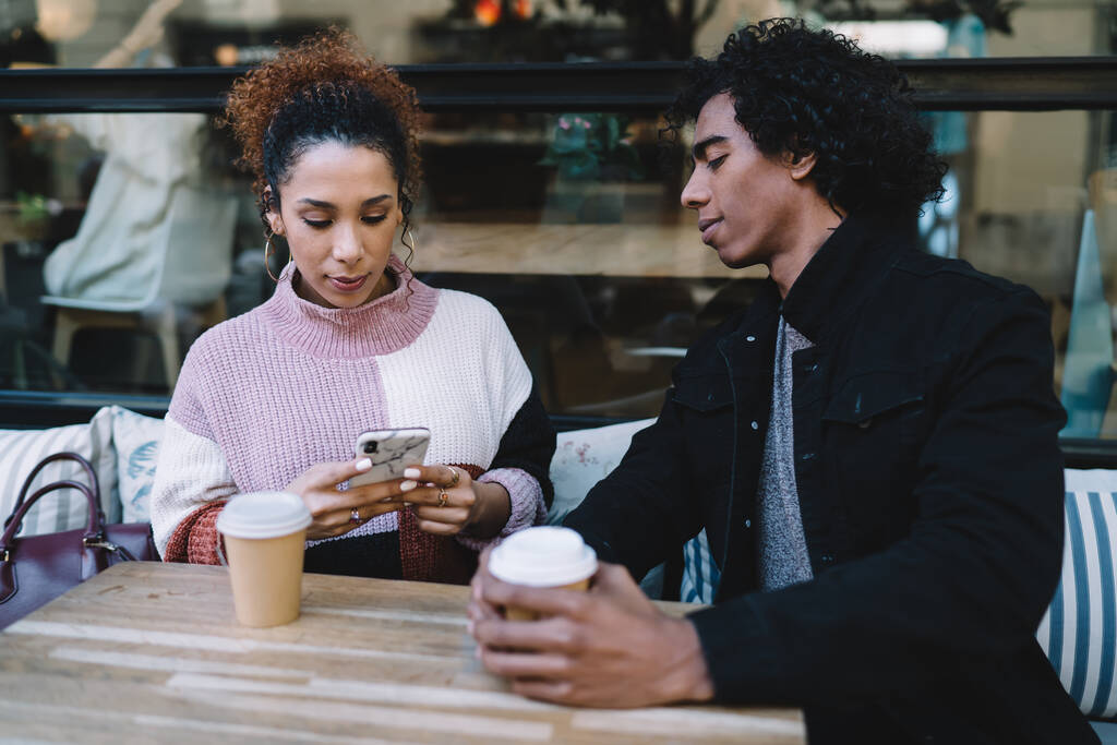 Ποικίλες νεαρός άνδρας με μαύρο σακάκι κοιτάζοντας γυναίκα φίλο σε casual πουλόβερ χρησιμοποιώντας το κινητό, ενώ πίνοντας καφέ στο σύγχρονο καφέ - Φωτογραφία, εικόνα
