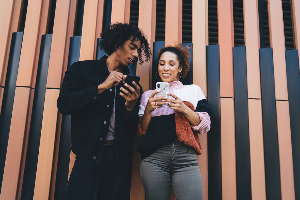 Alatta a figyelmes fekete férfi és mosolygós nő alkalmi ruhás olvasás üzeneteket okostelefonok, miközben hűvös együtt hétvégén - Fotó, kép