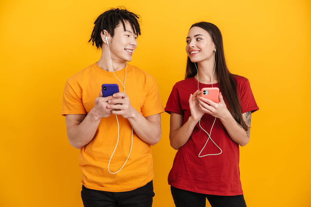 Młoda wielorasowa para słuchająca muzyki ze słuchawkami i komórkami odizolowanymi nad żółtą ścianą - Zdjęcie, obraz