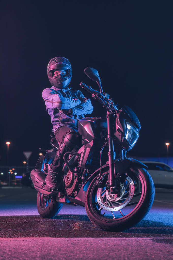 Мотоцикліст сидить на мотоциклі в неоновому світлі на порожній парковці вночі
 - Фото, зображення