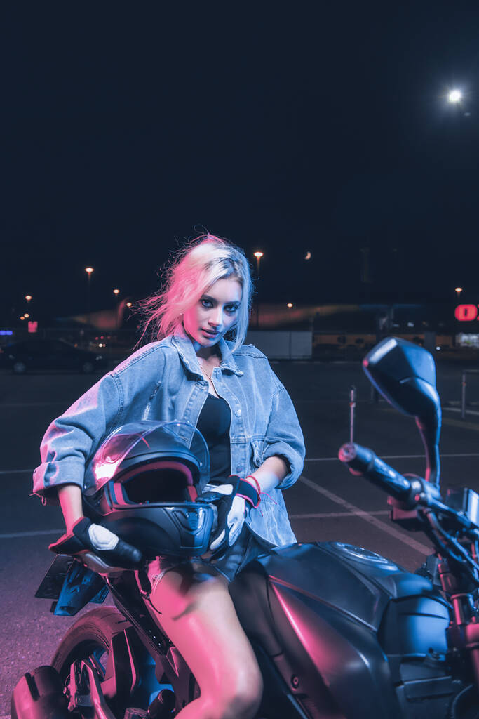 retrato de uma menina nos raios de luz de néon em uma moto à noite em um estacionamento vazio - Foto, Imagem
