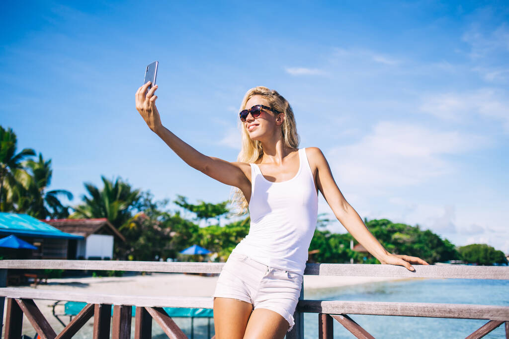 Щасливий кавказький турист у сонцезахисних окулярах з використанням передньої камери мобільного зв'язку для самостійного медіа-контенту під час відпусток в Індонезії, весела молода жінка знімає відео і посміхається - Фото, зображення