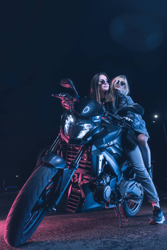 un par de chicas enamoradas en una moto en los rayos de luz de neón en un estacionamiento vacío por la noche - Foto, imagen