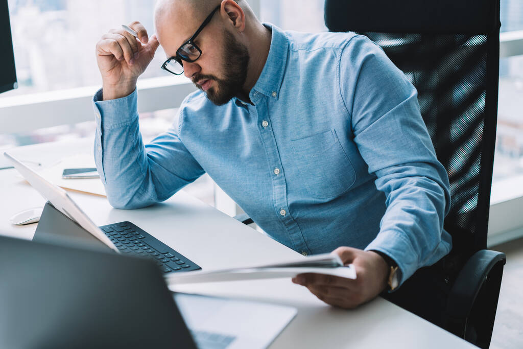 Soustředěný vážně vousatý muž zaměstnanec nosí formální oblečení a brýle pracuje na tabletu, zatímco sedí za stolem ve světle moderní kanceláře - Fotografie, Obrázek