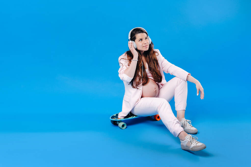 беременная женщина в розовом костюме сидит на скейтборде в наушниках и слушает музыку на синем фоне. - Фото, изображение