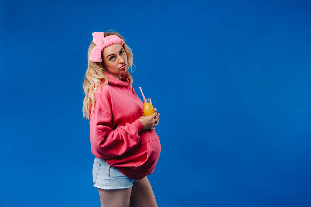 fille enceinte en vêtements roses avec une bouteille de jus sur un fond bleu. - Photo, image