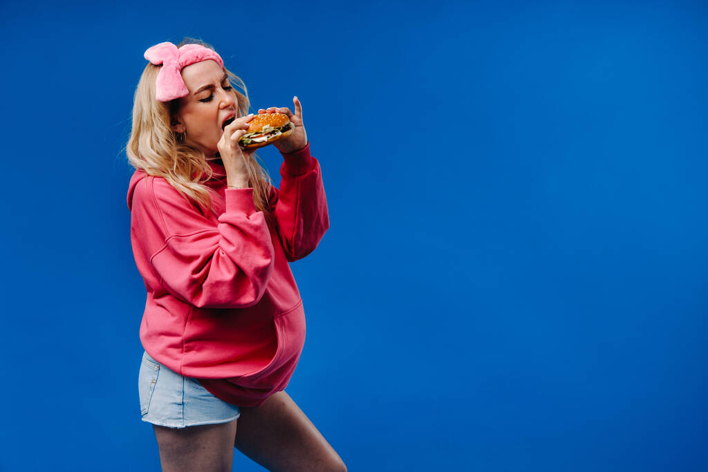 une fille enceinte en vêtements roses avec un hamburger dans ses mains sur un fond bleu. - Photo, image
