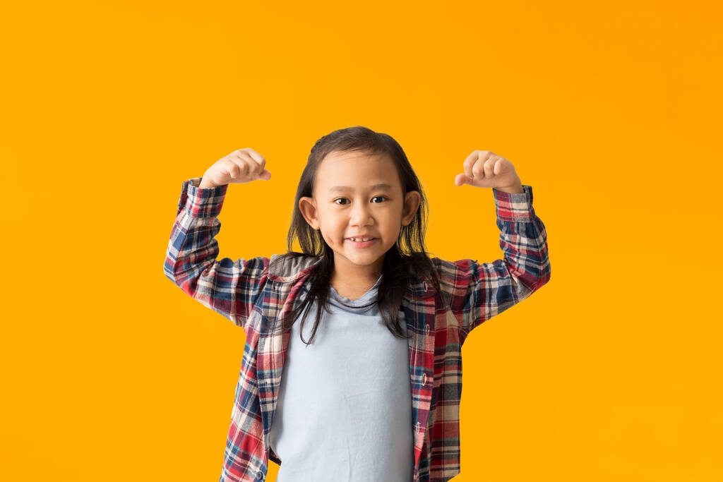 Pozytywne azjatyckie dziecko dziewczynka podnosi ramiona pokazuje mięśnie udaje bardzo silne i potężne uśmiechy, izolowane na żółtym tle  - Zdjęcie, obraz