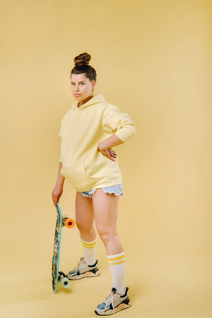 Вагітна дівчина в жовтій куртці зі скейтбордом в руках на жовтому тлі
. - Фото, зображення