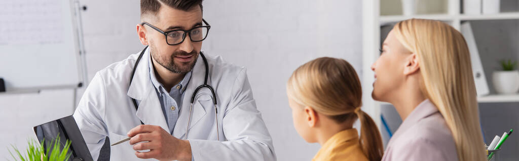Lääkäri katselee lasta osoittaessaan läppäriä lähellä hymyilevää vanhempaa, banneri  - Valokuva, kuva