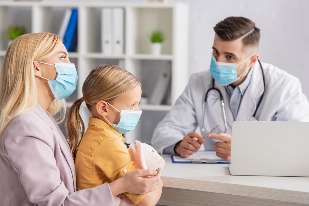 Mãe e criança em máscaras médicas sentado perto de médico borrado e laptop  - Foto, Imagem