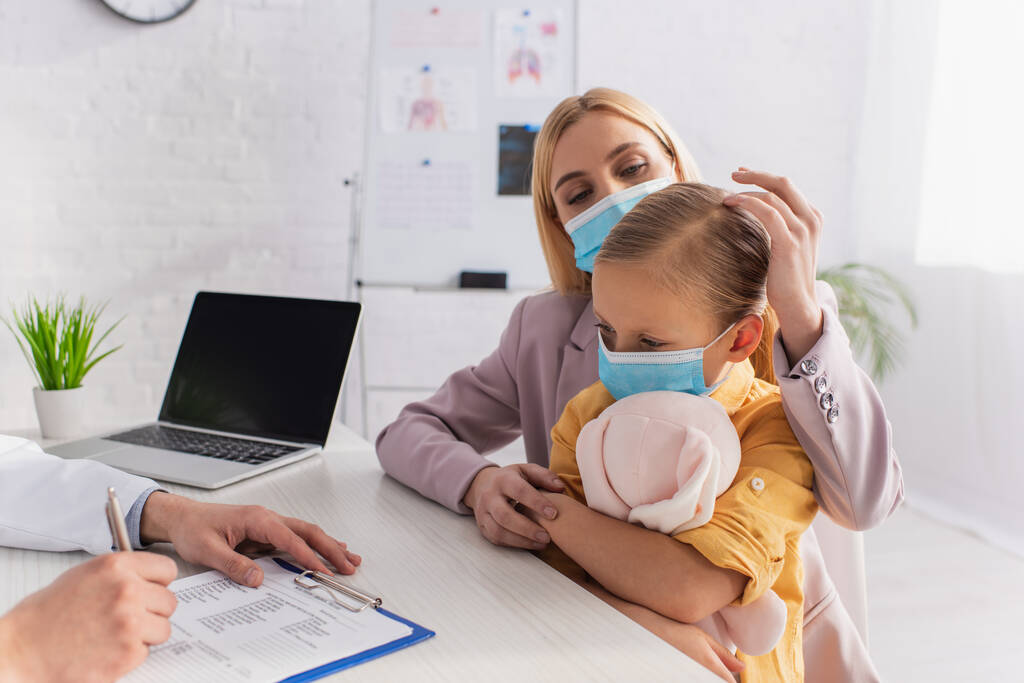 Μητέρα κοιτάζοντας την κόρη σε ιατρική μάσκα κοντά σε οικογενειακό γιατρό γράφοντας στο πρόχειρο  - Φωτογραφία, εικόνα