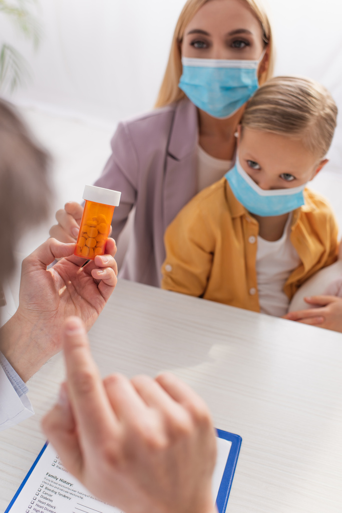 Таблетки в руках размытого педиатра и матери с ребенком в медицинских масках  - Фото, изображение